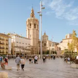 Gente pasea por la plaza frente a la Catedral de Valencia, con el icónico campanario del Miguelete dominando el cielo azul.