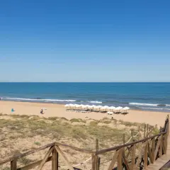 Pasarela de madera hacia la playa de Miramar, con sombrillas blancas alineadas y el mar azul bajo un cielo despejado.