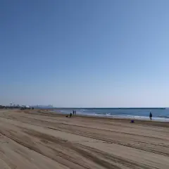 Amplia y tranquila playa de Meliana con arena marcada por vehículos. El mar azul se extiende hasta el horizonte, con algunas personas paseando.
