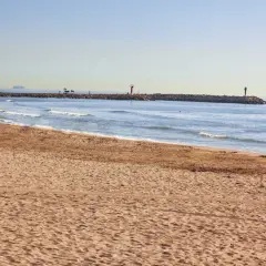 Arena dorada en la playa Port Saplaya, con el mar azul y un espigón con faros.