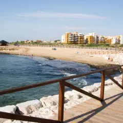 Paseo de madera junto al mar en la cala Piteras. Edificios modernos y playa de arena dorada.