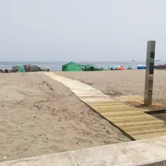 Pasarela de madera hacia las playas con duchas en Alicante. Gente disfrutando del sol bajo sombrillas y tiendas verdes.