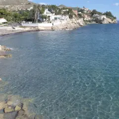 Aguas cristalinas bañan la costa rocosa de cala Piteres, con casas blancas y palmeras en la ladera.
