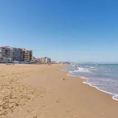 Arena dorada y mar azul en la playa La Roqueta, con edificios de apartamentos y montañas al fondo.