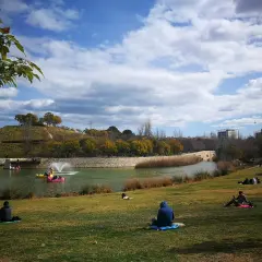 Gente relajándose en el césped del parque de cabecera, con un lago y barcas de pedales al fondo.