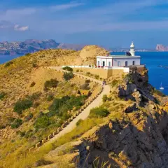 Faro blanco en acantilado rocoso del parque natural de la Serra Gelada. Camino serpenteante, mar azul y montañas al fondo.