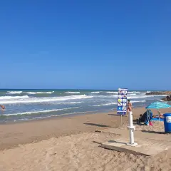 Un día soleado en la playa el Marenyet. Olas rompen en la orilla mientras una persona se baña. Hay un cartel de normas y una sombrilla.