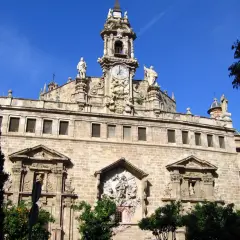 La **iglesia de los Santos Juanes**, con su imponente fachada de piedra, torre con reloj y estatuas, se alza majestuosa bajo un cielo azul intenso.