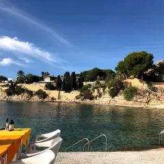 Mesa con mantel naranja y sillas blancas junto al mar cristalino en cala les Bassetes. Casas y vegetación en la ladera.