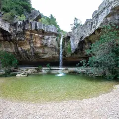 Cascada cayendo en **charcos de quesa** de agua cristalina, rodeada de rocas y vegetación exuberante.
