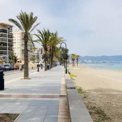 Paseo marítimo con palmeras y edificios modernos junto a la playa del Gurugú. El mar azul y las montañas al fondo crean un paisaje sereno.