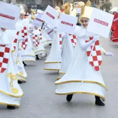 Participantes en la cabalgata del ninot, con trajes blancos y rojos, portan carteles con palabras como "Ciencia" y "Educació".