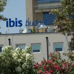El cartel azul "ibis budget" destaca sobre el edificio del hotel Ibis Budget Alicante, rodeado de exuberante vegetación y flores rosadas y blancas.