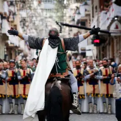 Un jinete con capa blanca lidera un desfile de moros y cristianos, con gente observando y banderas en las calles.