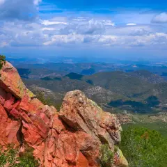 Rocas rojizas y vegetación verde en primer plano, con un vasto paisaje montañoso de la sierra de Espadán bajo un cielo azul con nubes.