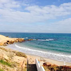 Escaleras descienden a la playa rocamar, con arena dorada, rocas y mar azul bajo un cielo nublado.