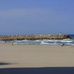 Una playa tranquila con una rompeolas de rocas. Gente relajándose en sillas de playa y paseando. El mar azul y el cielo despejado crean una escena de verano perfecta.