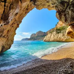 Vista desde una cueva hacia la cala Testos en Benitachell. Aguas turquesas, arena de guijarros y acantilados dorados bajo un cielo azul.