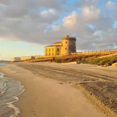 Atardecer en la playa del Conde, con una torre antigua y un paseo de madera. El mar acaricia la arena dorada bajo un cielo nublado.