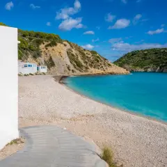 Casa blanca con puerta azul en la playa de guijarros de Vall d'Ebo, con aguas turquesas y colinas verdes.