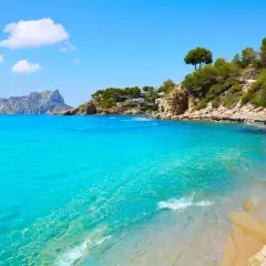 Aguas turquesas y arena dorada en la cala Pinets, con rocas y pinos coronando la costa. Un paraíso mediterráneo.