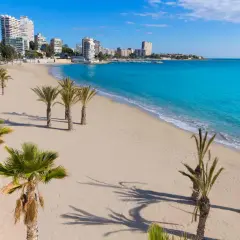 La **playa de Altea** se extiende con arena dorada y palmeras. El mar Mediterráneo, de un azul turquesa intenso, baña la costa, mientras edificios modernos se alzan al fondo.