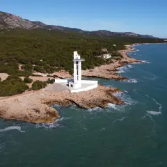Faro blanco moderno en la costa rocosa de la Sierra de Irta, con el mar azul y montañas verdes al fondo.