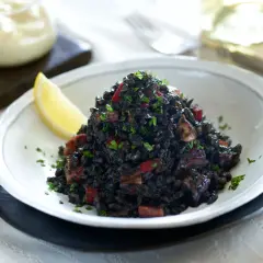 Delicioso arroz negro con trozos de calamar y pimiento rojo, adornado con perejil fresco y una rodaja de limón.