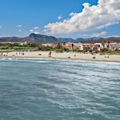 Olas azules rompen en la playa de arena de la playa Venecia Gandía, con gente disfrutando del sol y las montañas al fondo.