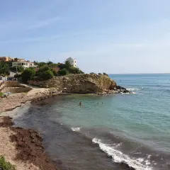 La cala Coveta, con su playa de arena y rocas, invita a disfrutar del mar Mediterráneo. Casas blancas coronan la colina.
