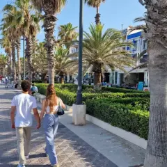 Pareja paseando por el paseo marítimo de Valencia, con palmeras, gente y edificios al fondo.