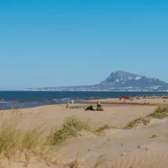 Dunas y mar en la playa de Rabdells, con gente disfrutando del sol y el paisaje montañoso al fondo.