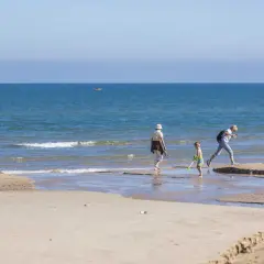 Un niño y dos adultos disfrutan de un día soleado en la playa Pau Pi, jugando cerca de la orilla del mar azul.