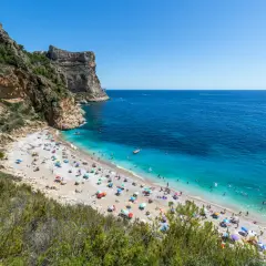 La **cala Moraig** es un paraíso de guijarros y aguas turquesas, con acantilados imponentes y gente disfrutando del sol bajo sombrillas de colores.