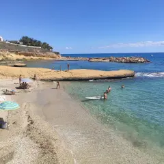 Gente disfrutando del sol y el mar en la cala Almadraba, Campello. Aguas cristalinas y rocas emergiendo del azul.