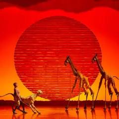 Escena de teatro en Valencia con un guepardo y jirafas con zancos ante un sol naranja.