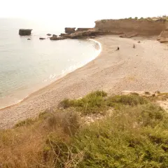 Una tranquila cala foradada con aguas cristalinas y rocas emergiendo del mar. Dos personas disfrutan de la soledad en la playa de guijarros.