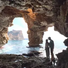 Pareja contemplando el mar desde la Cova dels Arcs, con sus impresionantes formaciones rocosas y el sol filtrándose.