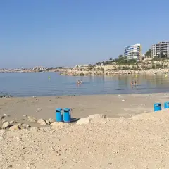 Día soleado en la cala Morro Blanc. Gente disfrutando del mar y la playa, con edificios modernos al fondo.