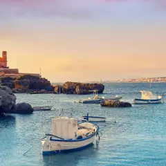 Barcas tranquilas en el puerto Santa Pola, con edificios históricos y la costa al fondo bajo un cielo anaranjado.
