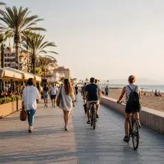 Gente pasea y monta en bicicleta por el paseo marítimo de la playa de la Patacona al atardecer, con palmeras y edificios al fondo.