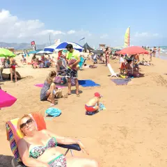 Día soleado en la playa de l'Ahuir, con gente disfrutando bajo sombrillas y el mar al fondo.