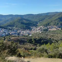 Vista panorámica de un pueblo en un valle rodeado de montañas boscosas. Se distingue la ermita de Sant Ramon.