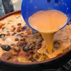 Un líquido naranja se vierte de un recipiente azul a una paella con carne y marisco.