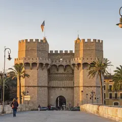 Las **Torres de Serranos**, imponentes puertas de la antigua muralla de Valencia, se alzan majestuosas bajo un cielo despejado. Palmeras y farolas flanquean el camino.