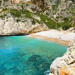 Aguas turquesas bañan la cala testos, una playa de guijarros rodeada de acantilados rocosos y vegetación.