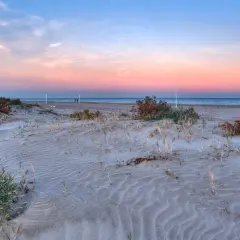 Dunas de arena con vegetación escasa en la **playa Almarda** al atardecer, con el mar y un cielo rosado de fondo.