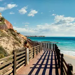 Paseo de madera con vistas a las mejores playas de Cabo Roig. Mar turquesa, arena dorada y un edificio amarillo en la ladera.