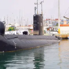 Un submarino negro, el Delfín, descansa en el puerto de Torrevieja, con mástiles de veleros y edificios al fondo.