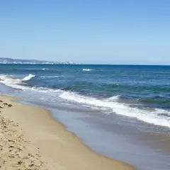 La playa de Xeraco, con arena dorada y el mar azul, se extiende hacia un horizonte con edificios y montañas.